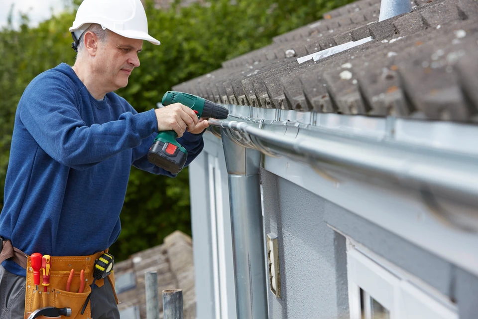 Ouvrier en bleu de travail avec casque blanc installe une gouttière en aluminium sur une maison résidentielle