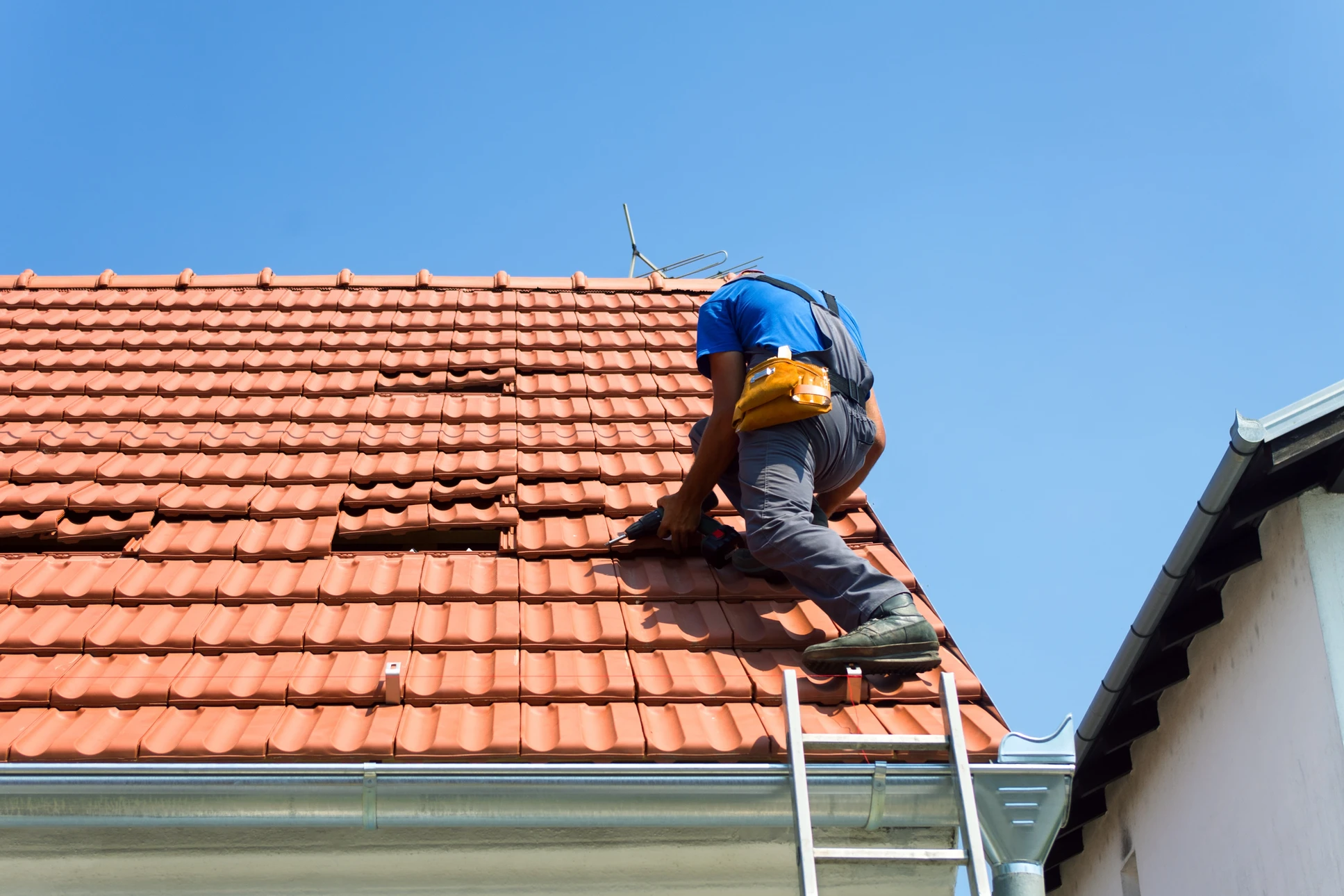 Ouvrier couvreur travaillant sur une toiture en tuiles rouges, portant casque de protection bleu et harnais de sécurité